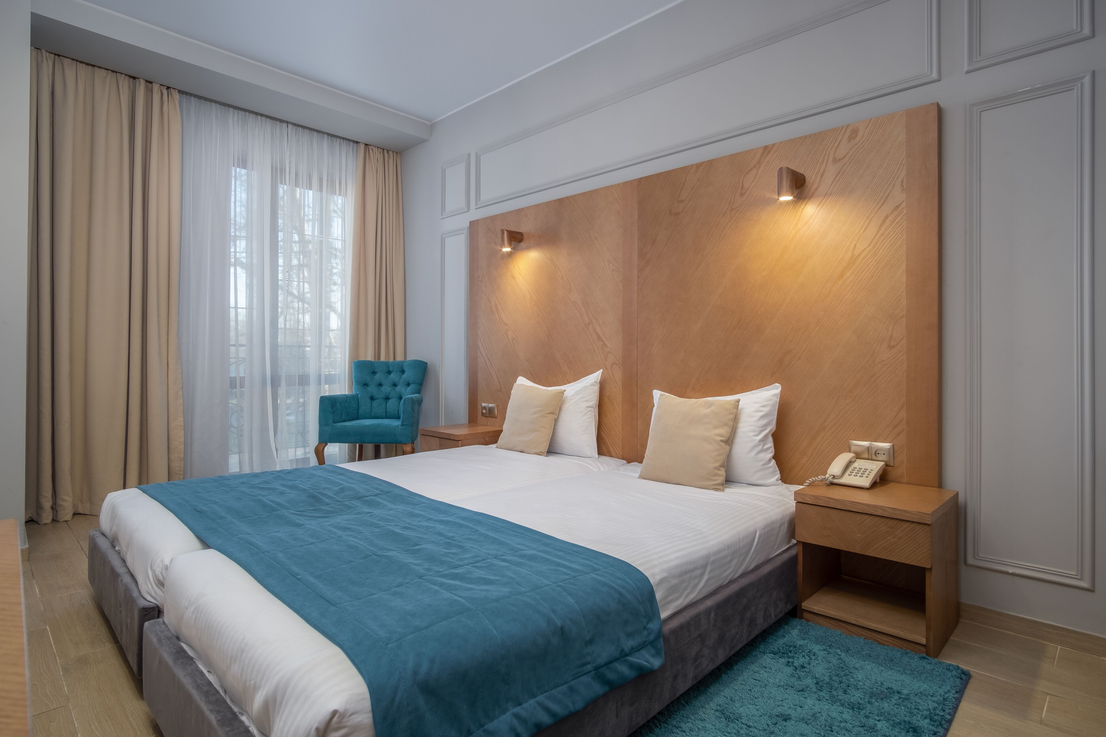 A modern hotel room with a large bed, a wooden headboard, a bedside phone, and a teal chair.