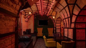 A cozy, dimly lit room with a vaulted ceiling, stone walls, and a flat-screen TV mounted on the wall. There are yellow chairs and a wooden table in the center.