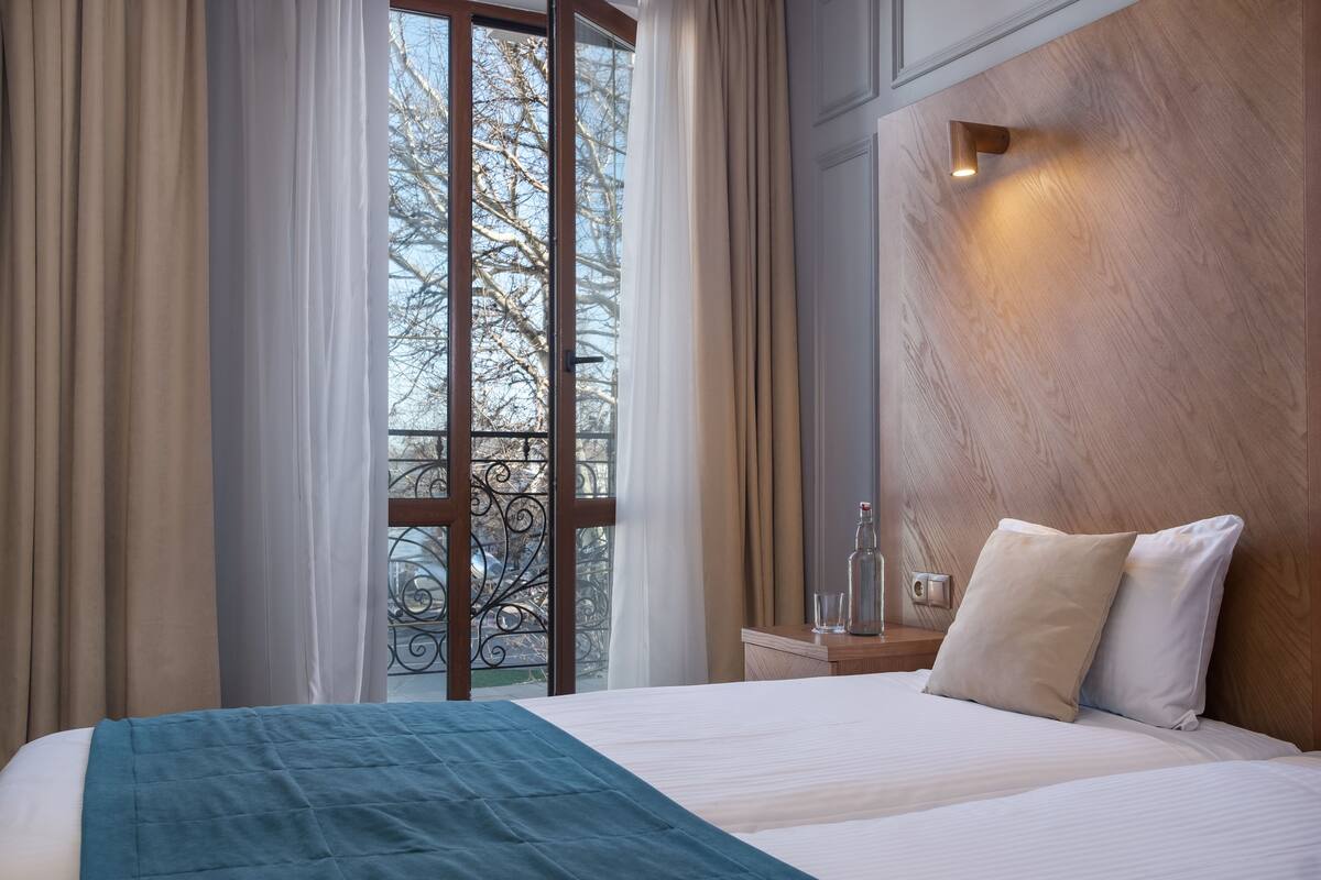 Hotelkamer met een groot bed, een houten hoofdbord, een nachtkastje met een glas en uitzicht op een balkon met metalen hekwerk.