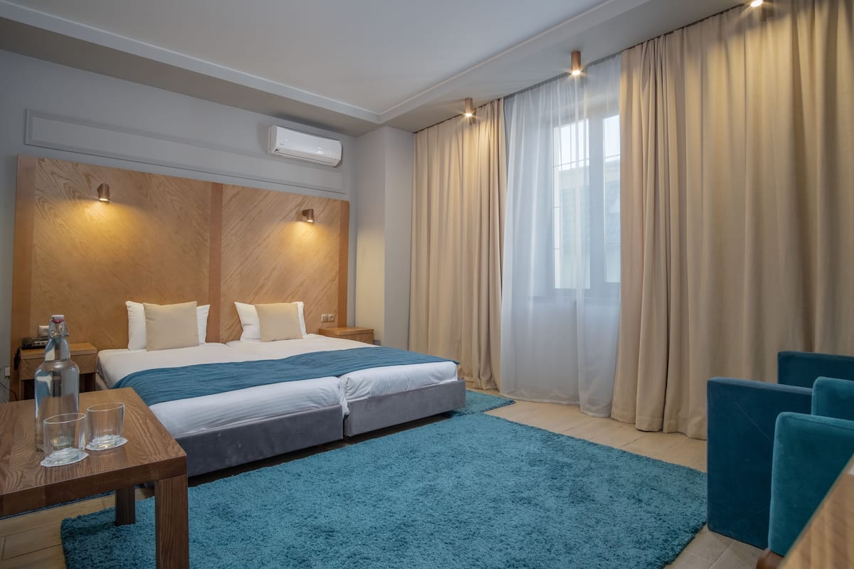 Een moderne hotelkamer met een groot bed, een houten hoofdbord, een blauw tapijt en uitzicht op de stad door dunne gordijnen.