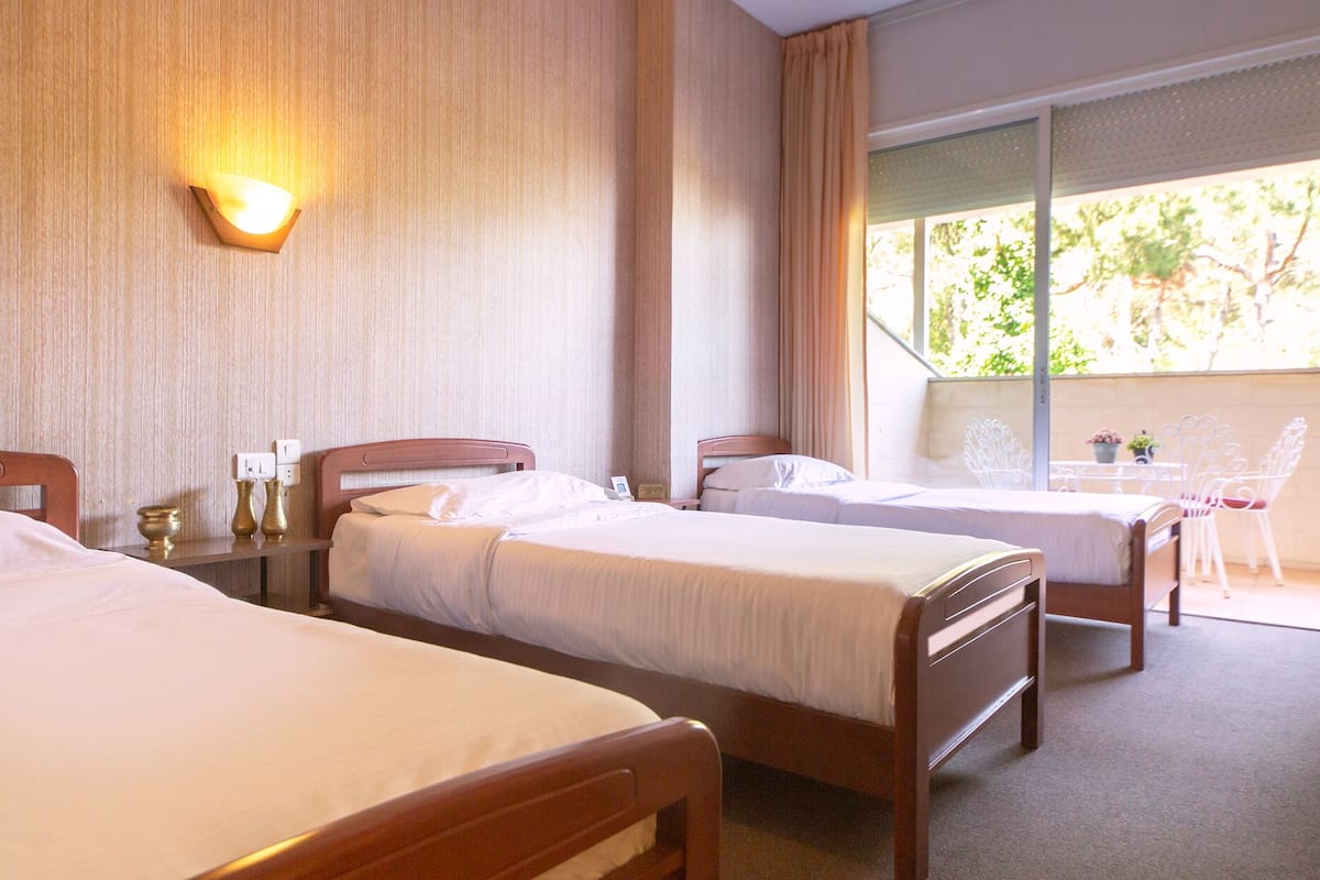 A hotel room with two single beds, a bedside table with a lamp, a window with curtains, and a balcony with outdoor furniture.