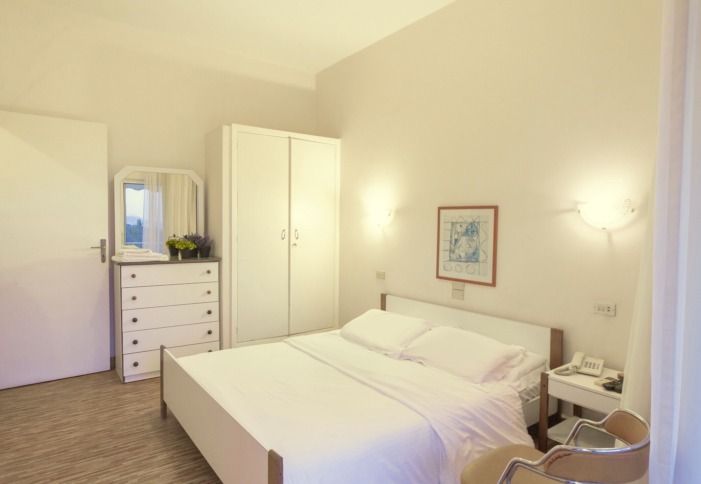 A hotel room with a bed, a dresser, a mirror, a bedside table with a phone, and a wall-mounted light fixture.