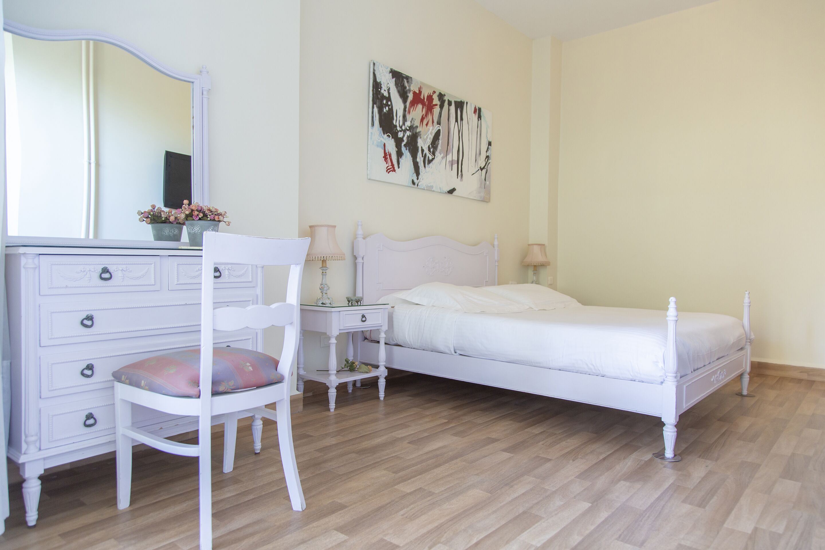 A bedroom with a white bed, a dresser, a chair, a bedside table, and a wall-mounted painting.