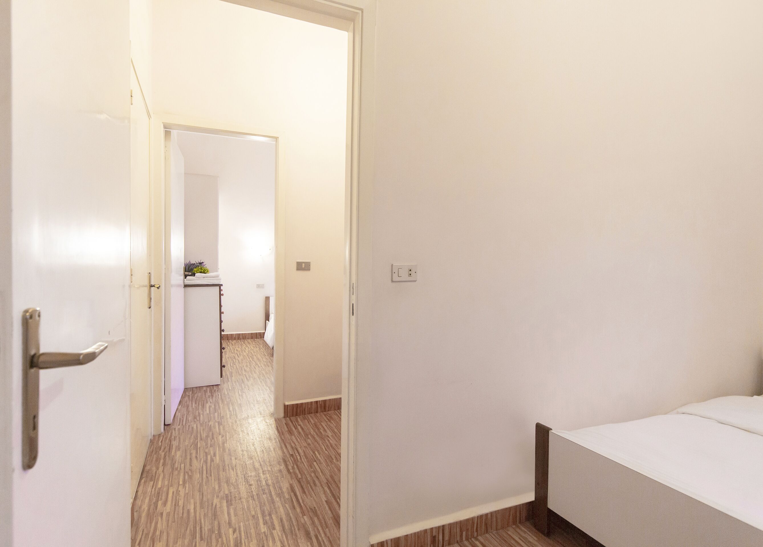 Une chambre d’hôtel avec un plancher en bois, un lit, un mur blanc et une porte donnant sur un couloir.