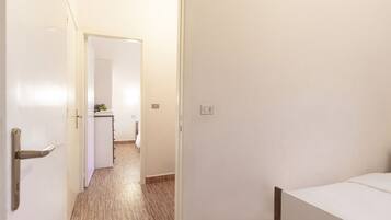 Une chambre d’hôtel avec un plancher en bois, un lit, un mur blanc et une porte donnant sur un couloir.