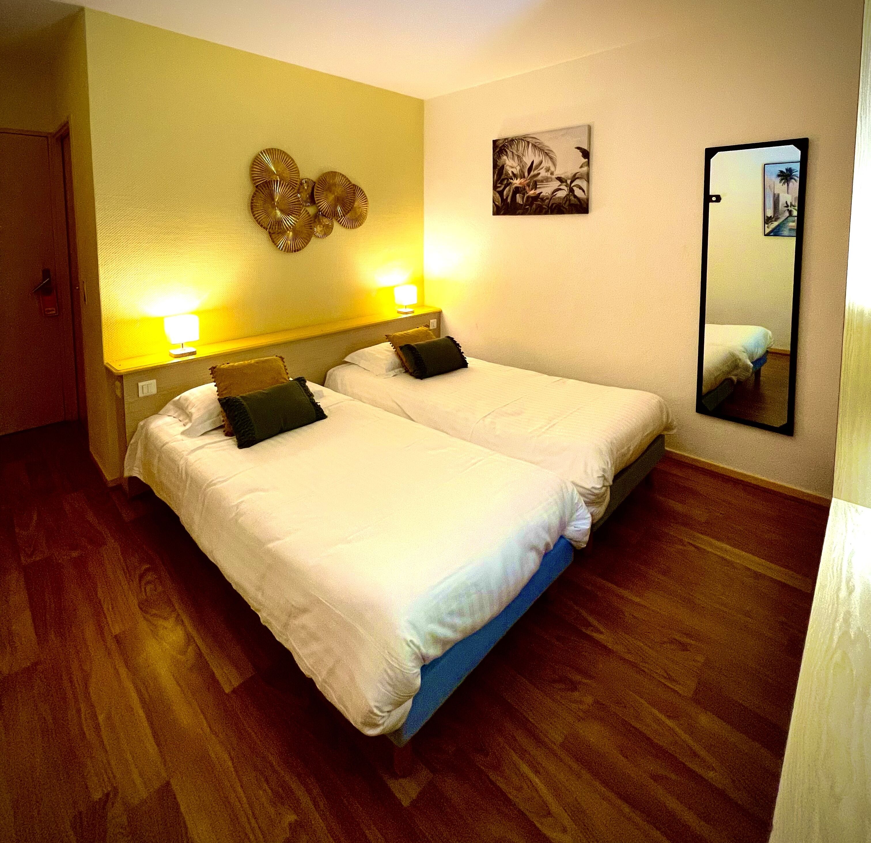 A hotel room with two beds, wooden flooring, a wall-mounted mirror, and a tropical-themed painting.