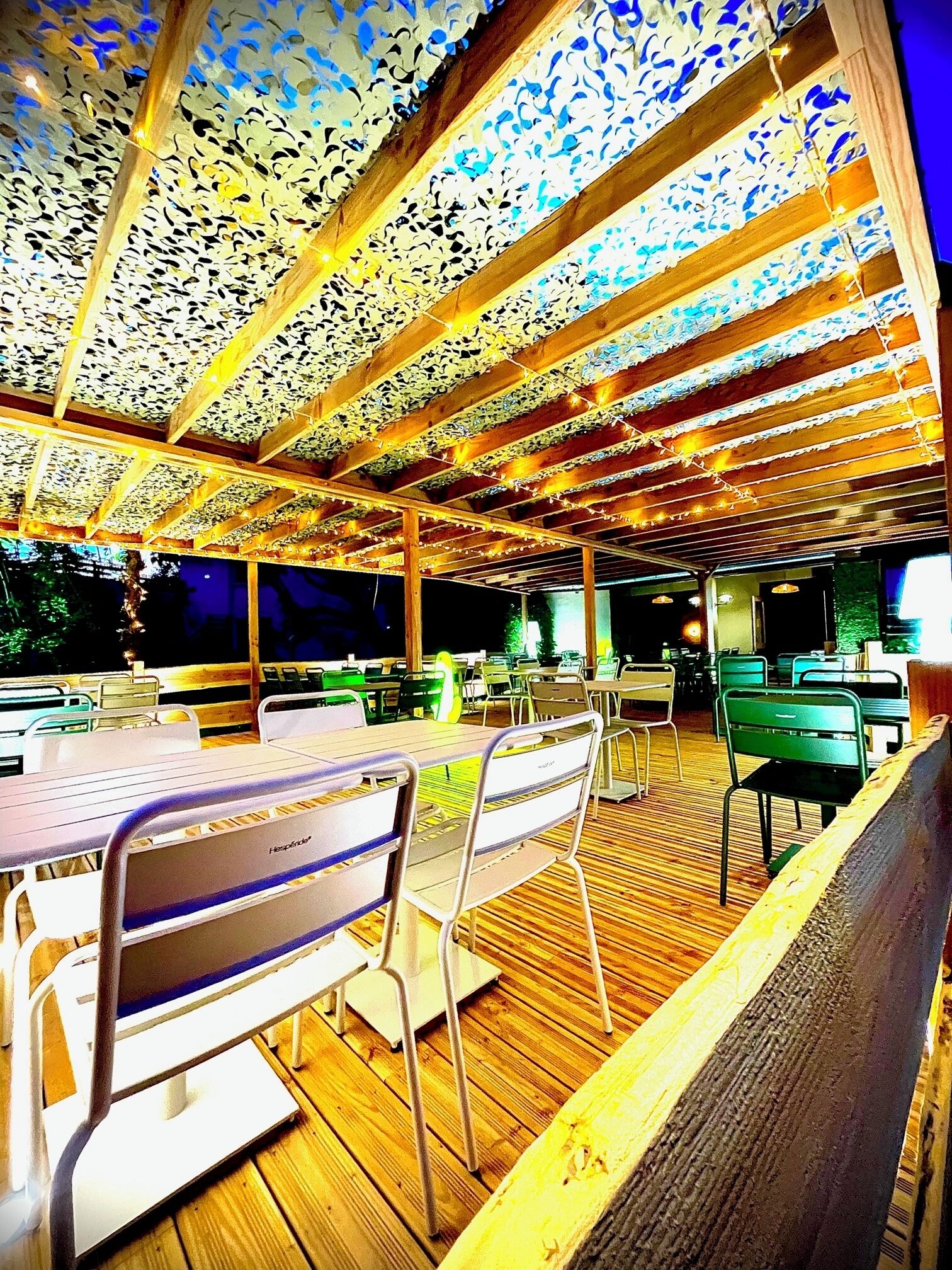 An outdoor dining area with wooden tables and chairs, a ceiling with a lace-like pattern, and string lights.