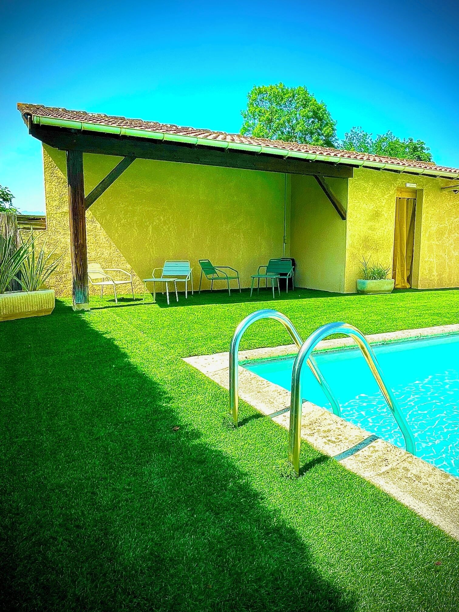 A pool area with a clear blue pool, a covered patio with chairs, and a small building with a door.