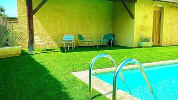 A pool area with a clear blue pool, a covered patio with chairs, and a small building with a door.