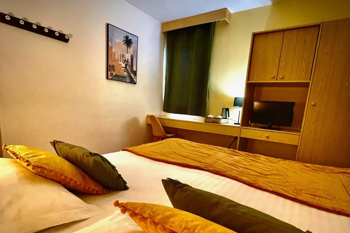 Une chambre d’hôtel avec deux lits, chacun étant équipé d’oreillers et d’une couverture. On y trouve un téléviseur posé sur une commode en bois et un tableau accroché au mur.