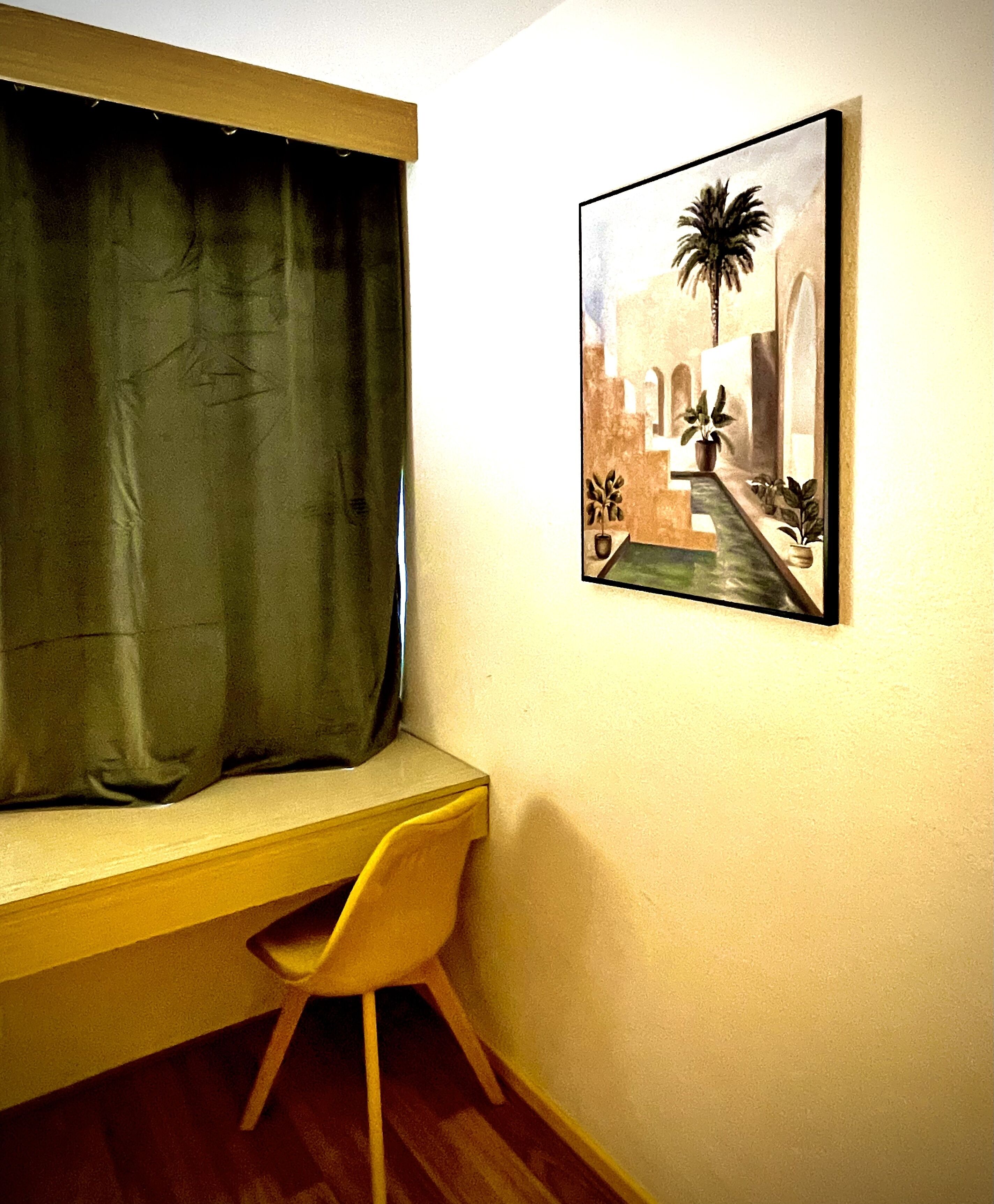 A room with a window, a desk, a chair, and a wall-mounted painting of a palm tree and architecture.