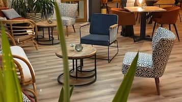 Un área de estar moderna con diversas opciones de asientos, incluyendo sofás, sillones y una mesa de comedor. Hay plantas en macetas y una puerta de salida visible.