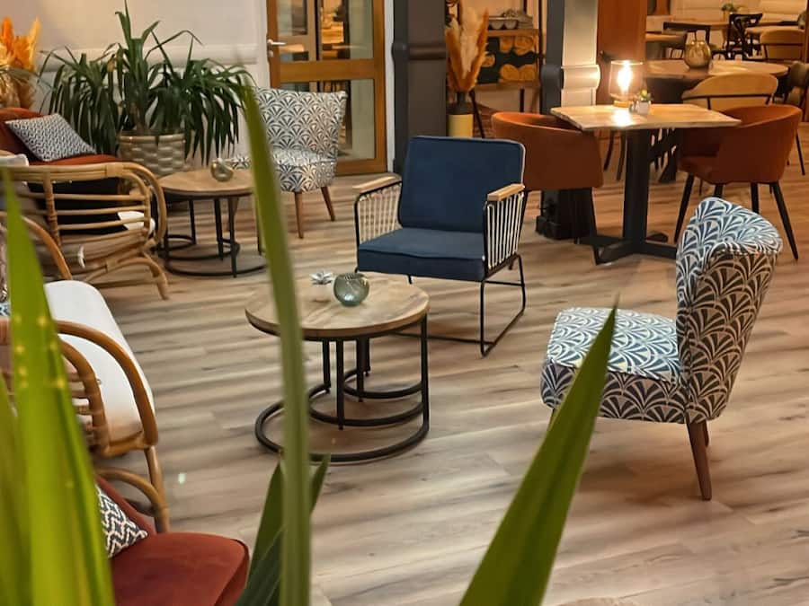 A modern lounge area with a variety of seating options including sofas, chairs, and a dining table. There are potted plants and a visible exit door.