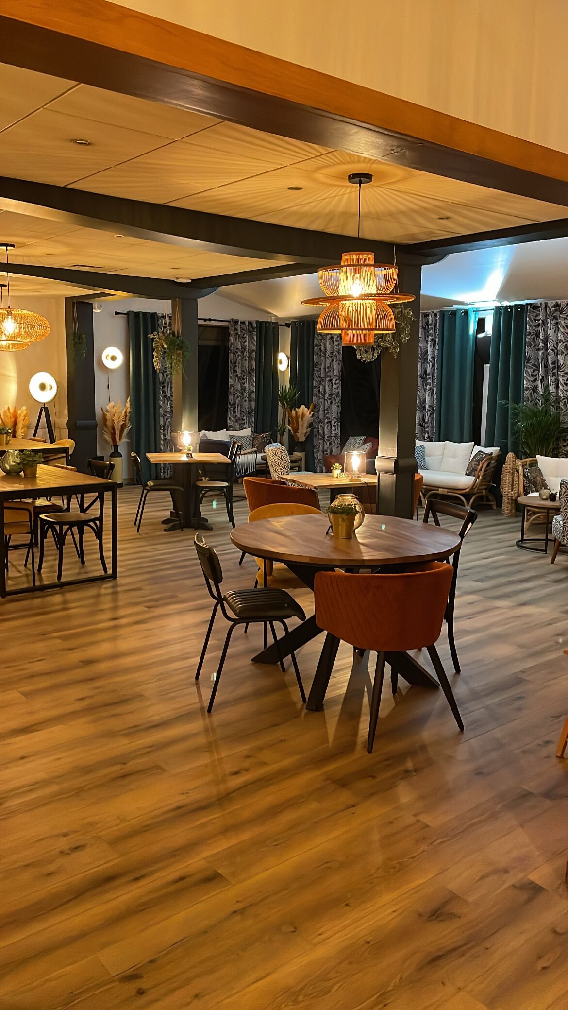 A spacious room with a wooden floor, a round dining table with chairs, and a large chandelier. There are curtains and a sofa in the background.