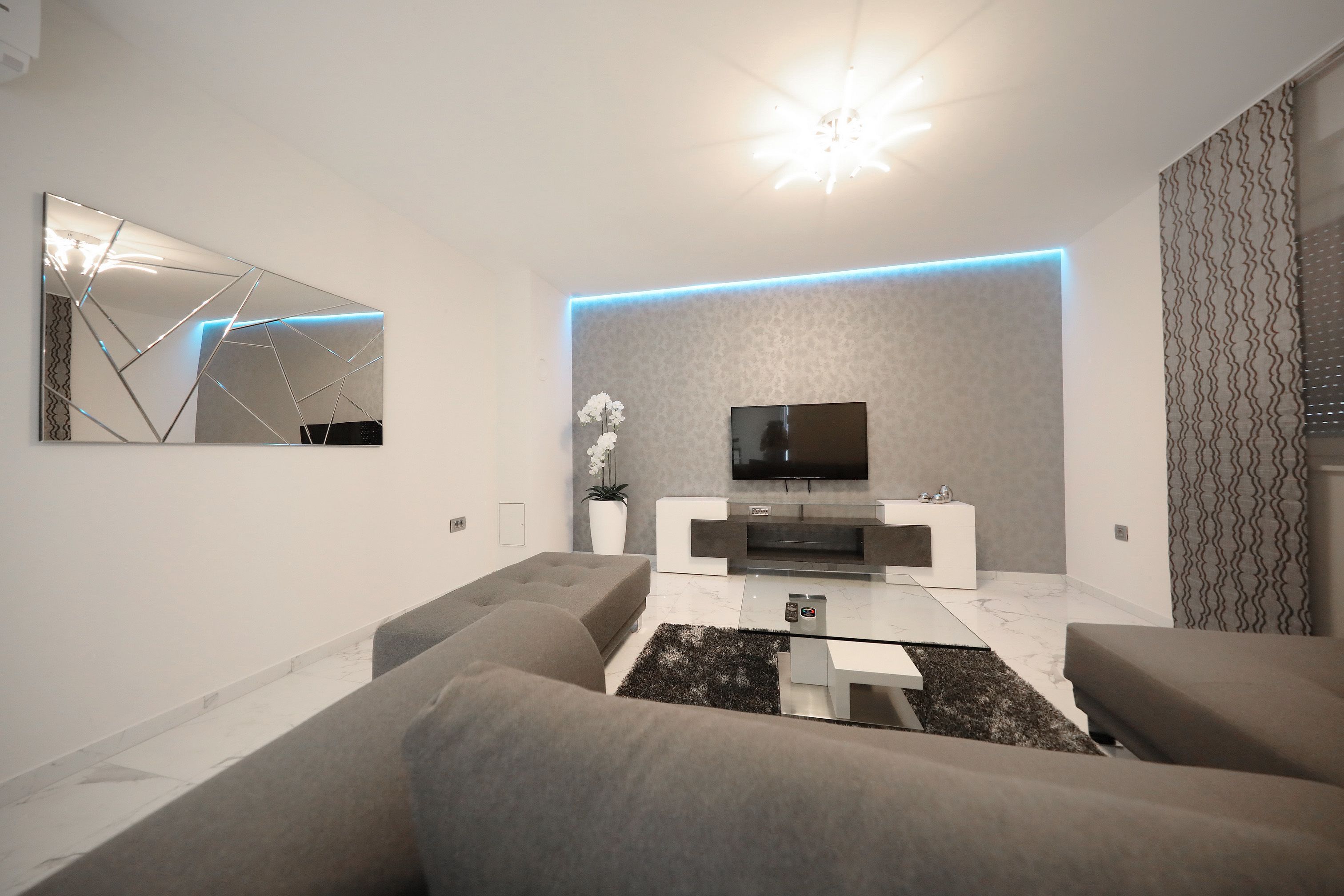 A modern living room with a flat-screen TV, grey sofas, a coffee table, and a patterned wall.