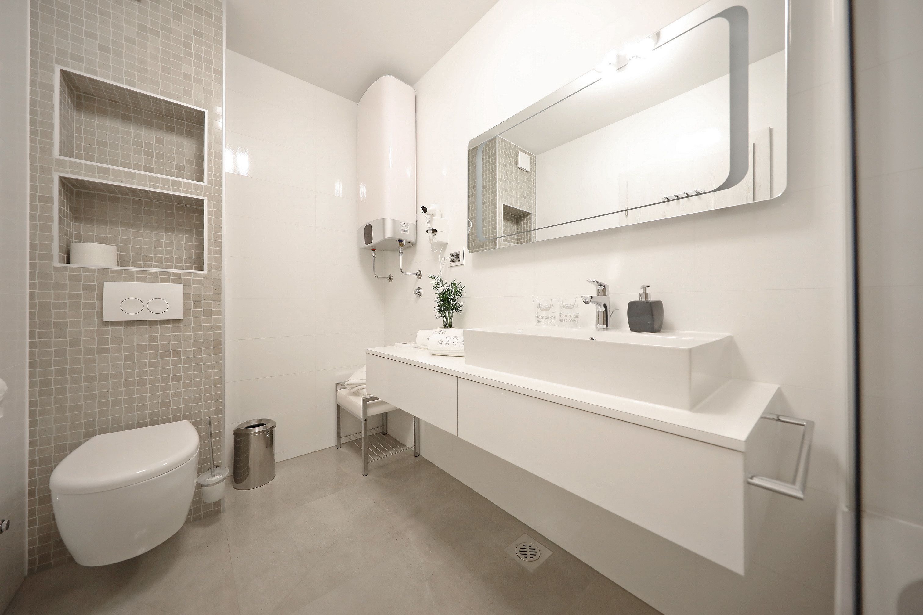 Une salle de bain moderne avec des toilettes blanches, un lavabo surmonté d’un miroir et une poubelle.