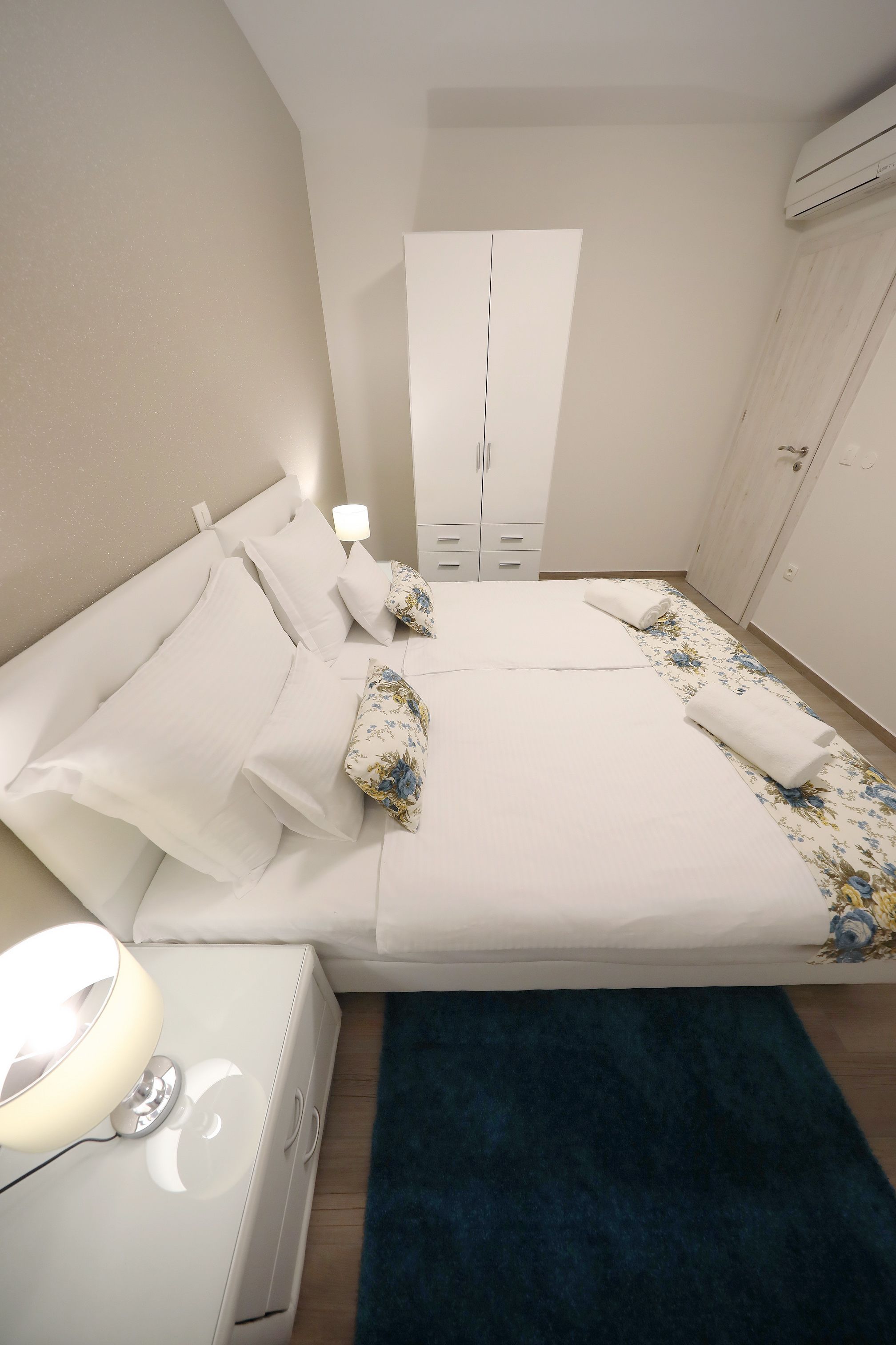 A neatly arranged bedroom with a bed, bedside tables, a wardrobe, and an air conditioning unit.