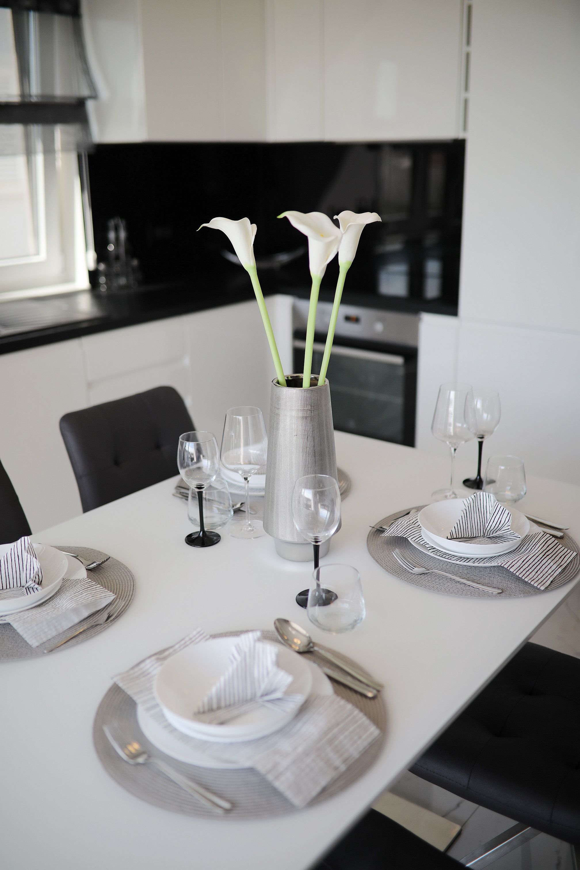 A modern dining table set with white plates, silverware, and clear wine glasses, accompanied by a vase with white flowers.