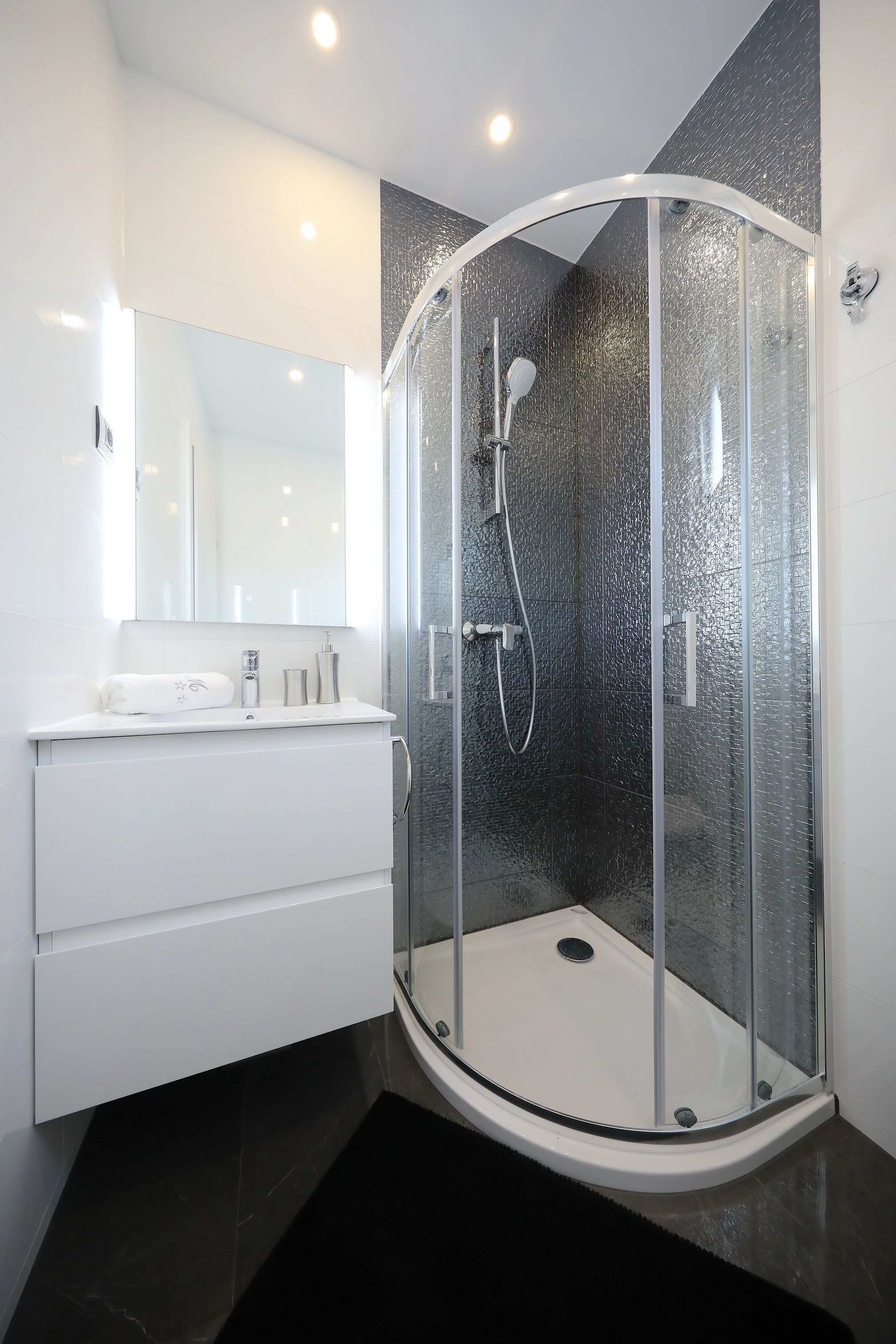 A modern bathroom with a glass-enclosed shower, a white vanity with a mirror, and dark tiled walls.