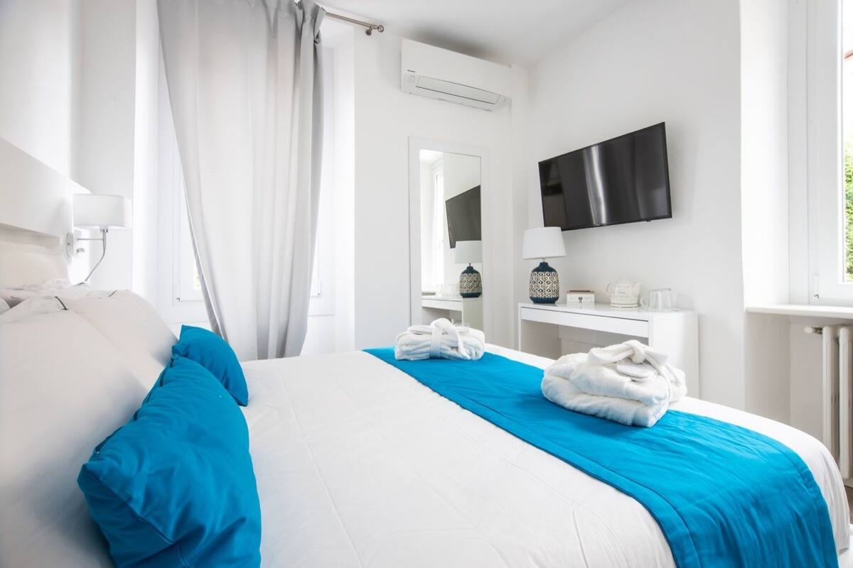 Habitación de hotel con una cama grande, un televisor de pantalla plana, una mesita de noche con lámpara y una ventana con cortinas.