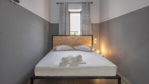 A neatly made bed with white linens and rolled towels, a headboard with a textured pattern, and a small bedside lamp. - Intra Muros Hostel (Heraklion)