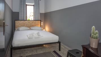 Una cama bien tendida con sábanas y almohadas blancas, una mesita de noche con un cactus, y vistas al exterior a través de una ventana con cortinas.