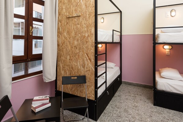 Una habitación con literas, una pequeña mesa con libros, una silla y una ventana con cortinas.