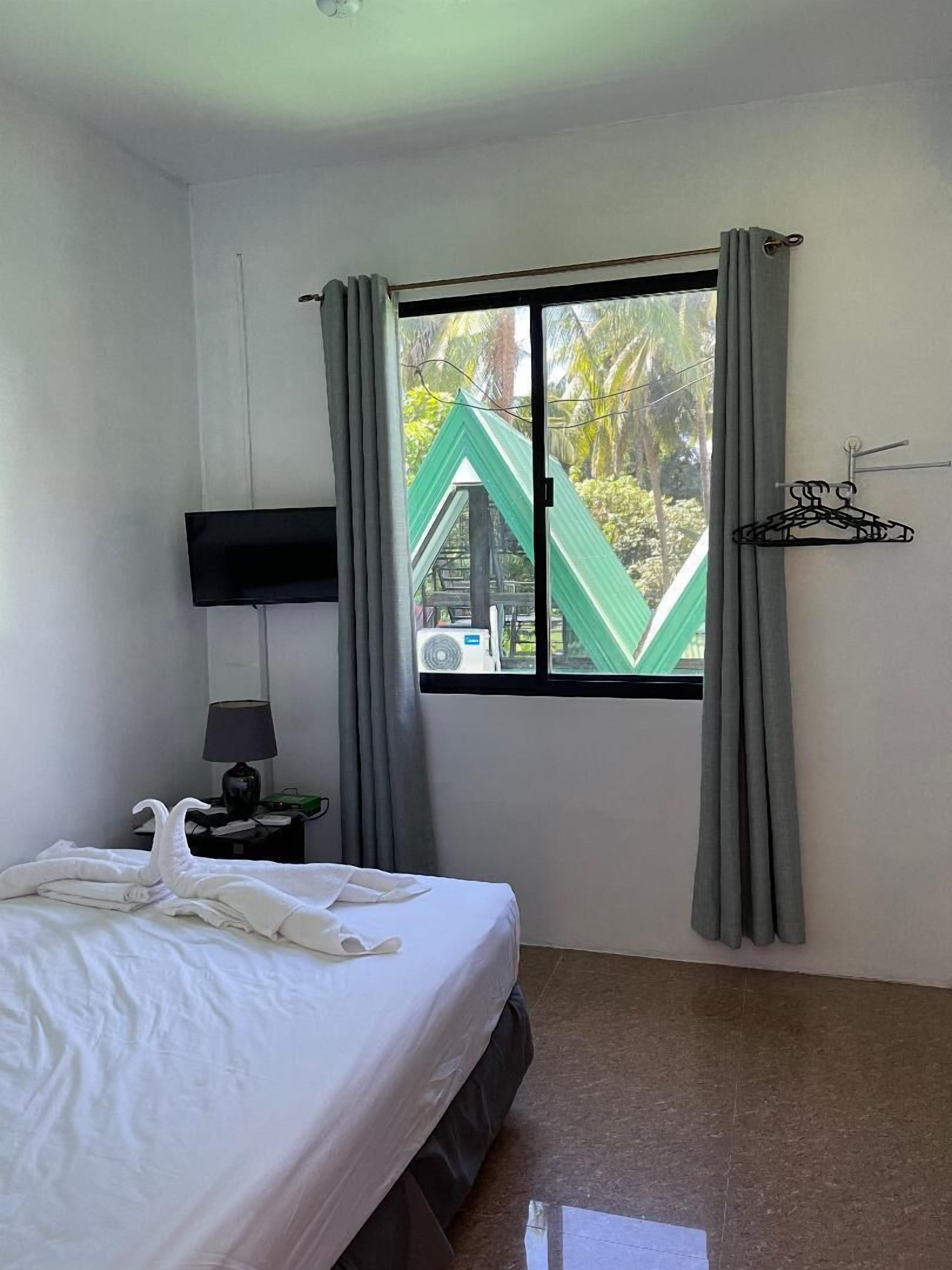 A hotel room with a bed, a nightstand with a lamp, a window with curtains, and a view of a green structure outside.