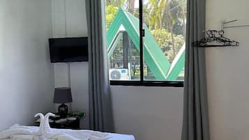 A hotel room with a bed, a nightstand with a lamp, a window with curtains, and a view of a green structure outside.