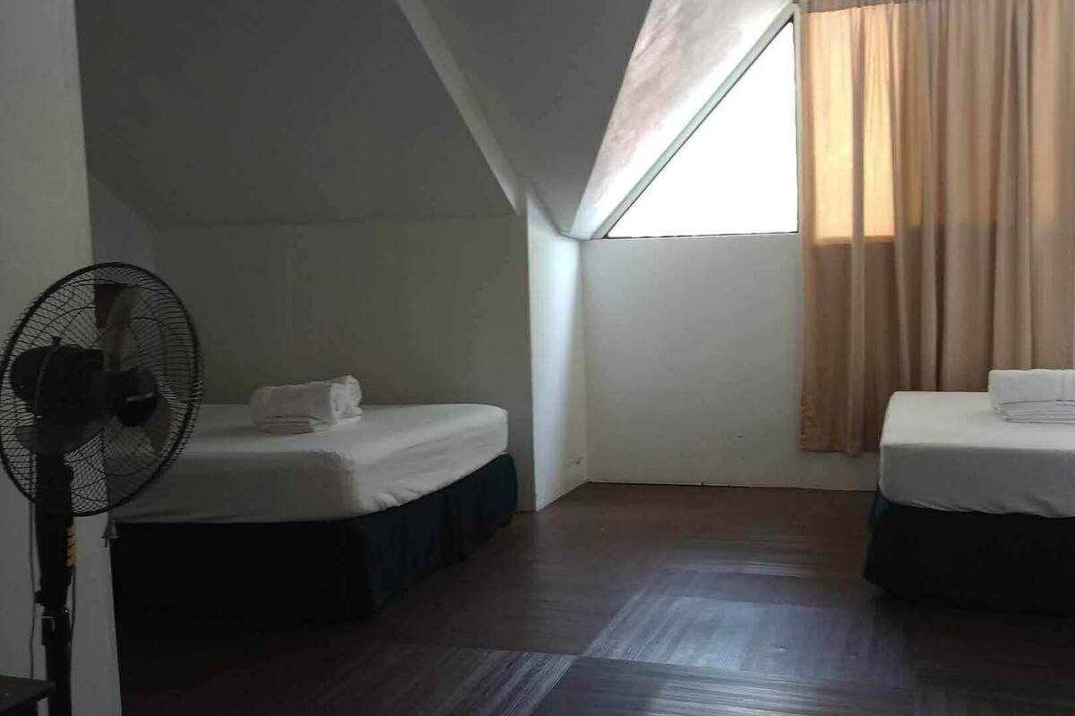 Una habitación pequeña y tenuemente iluminada con dos camas, un ventilador de pie y una ventana con cortinas.