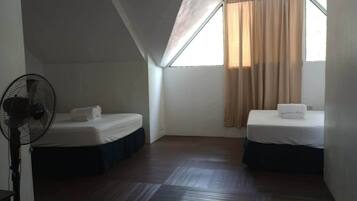 A small, dimly lit room with two beds, a standing fan, and a window with curtains.