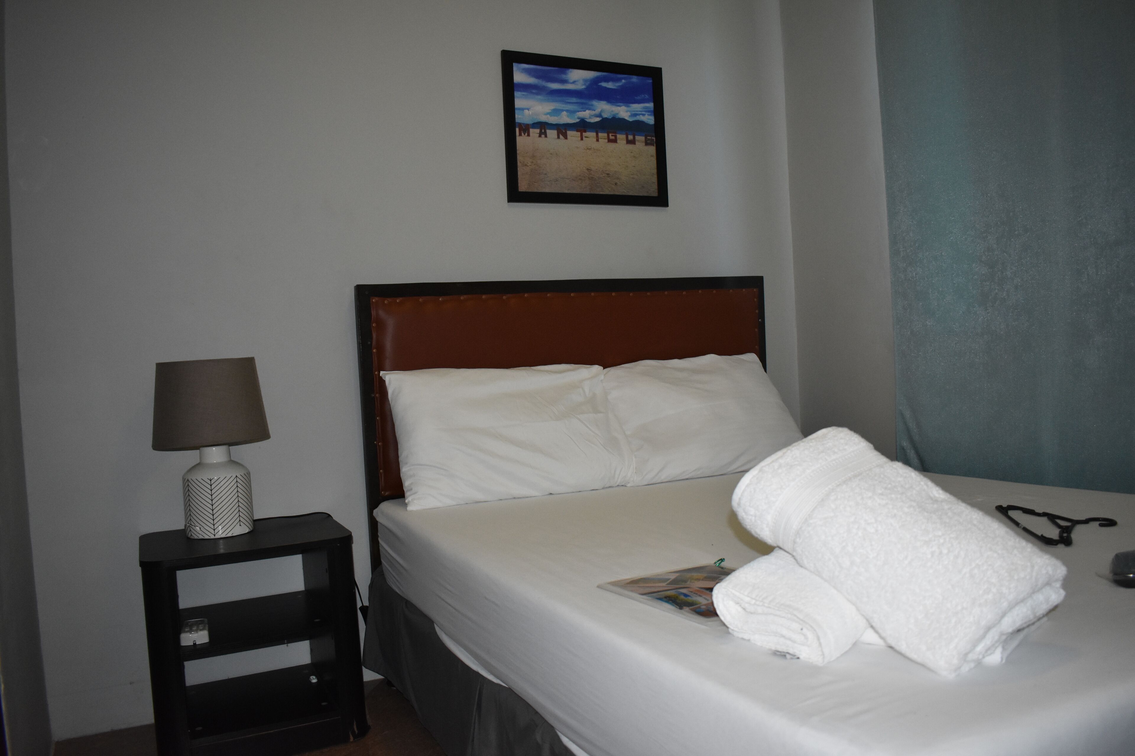 Una cama bien hecha con sábanas blancas, una toalla doblada y un par de gafas en la mesita de noche. Encima de la cama, un cuadro enmarcado adorna la pared, y a la izquierda se ve una mesita con una lámpara.