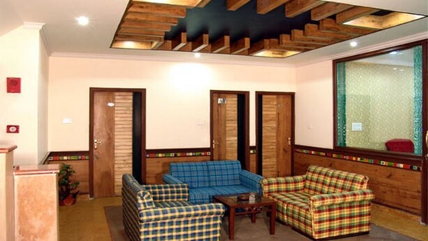 A lobby with wooden flooring, a blue sofa, a red sofa, and a wooden bench. There are two doors and a window with a green curtain. - Hotel Ivory Palace (New Delhi)