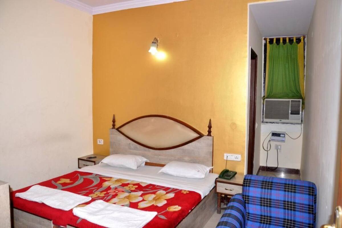 A hotel room with a bed, a bedside table, a nightstand, a wall-mounted air conditioner, and a window with a curtain.