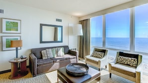 Habitación de hotel con un ventanal grande que ofrece vista al mar, un sofá, una silla y una mesita con bandeja.