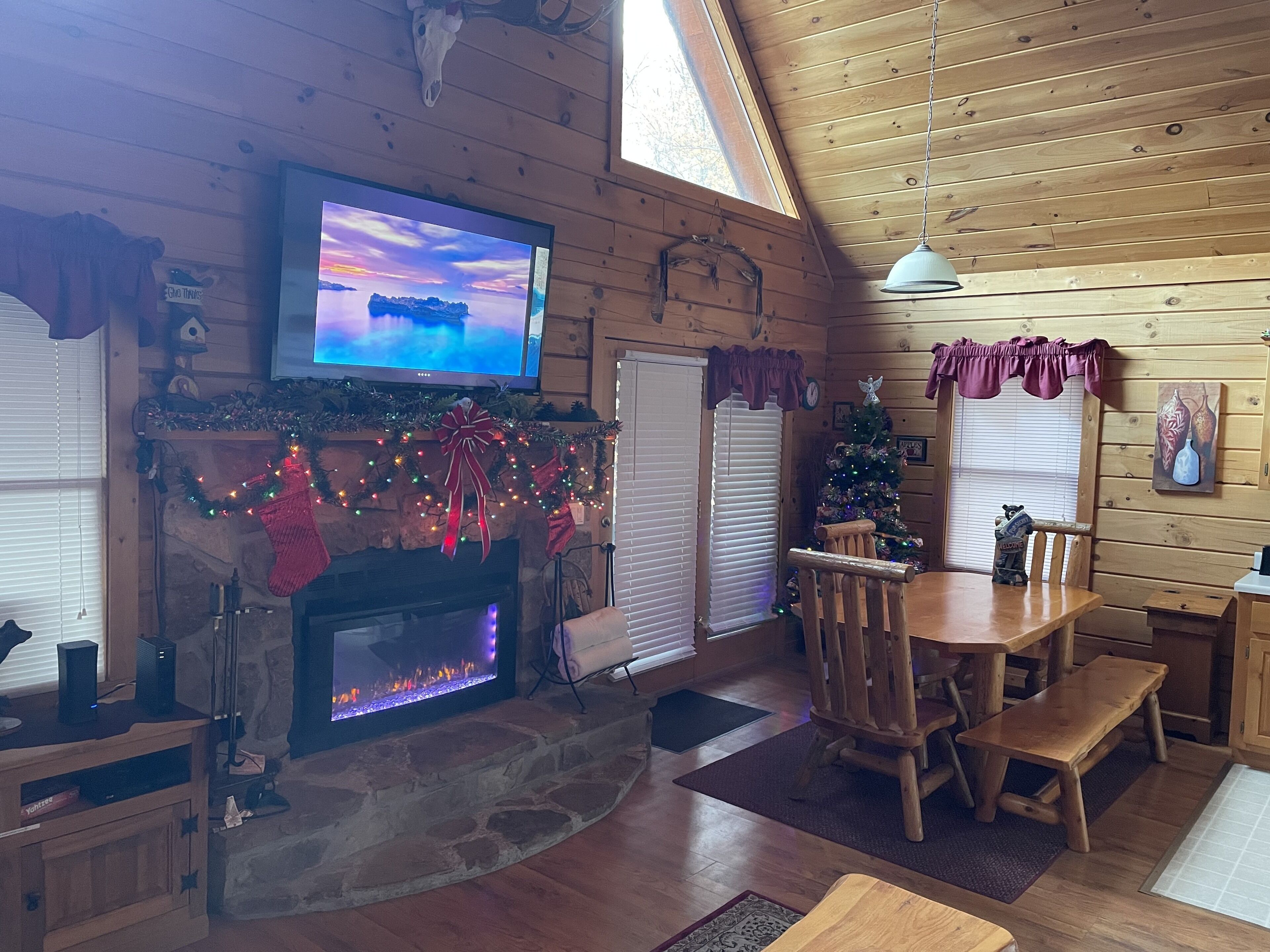 Un acogedor interior de cabaña con chimenea de piedra, mesa de comedor de madera y televisor montado en la pared.