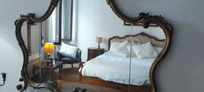 Une chambre à coucher avec un grand miroir décoratif, un lit recouvert de draps blancs, une table de chevet avec une lampe et une chaise.