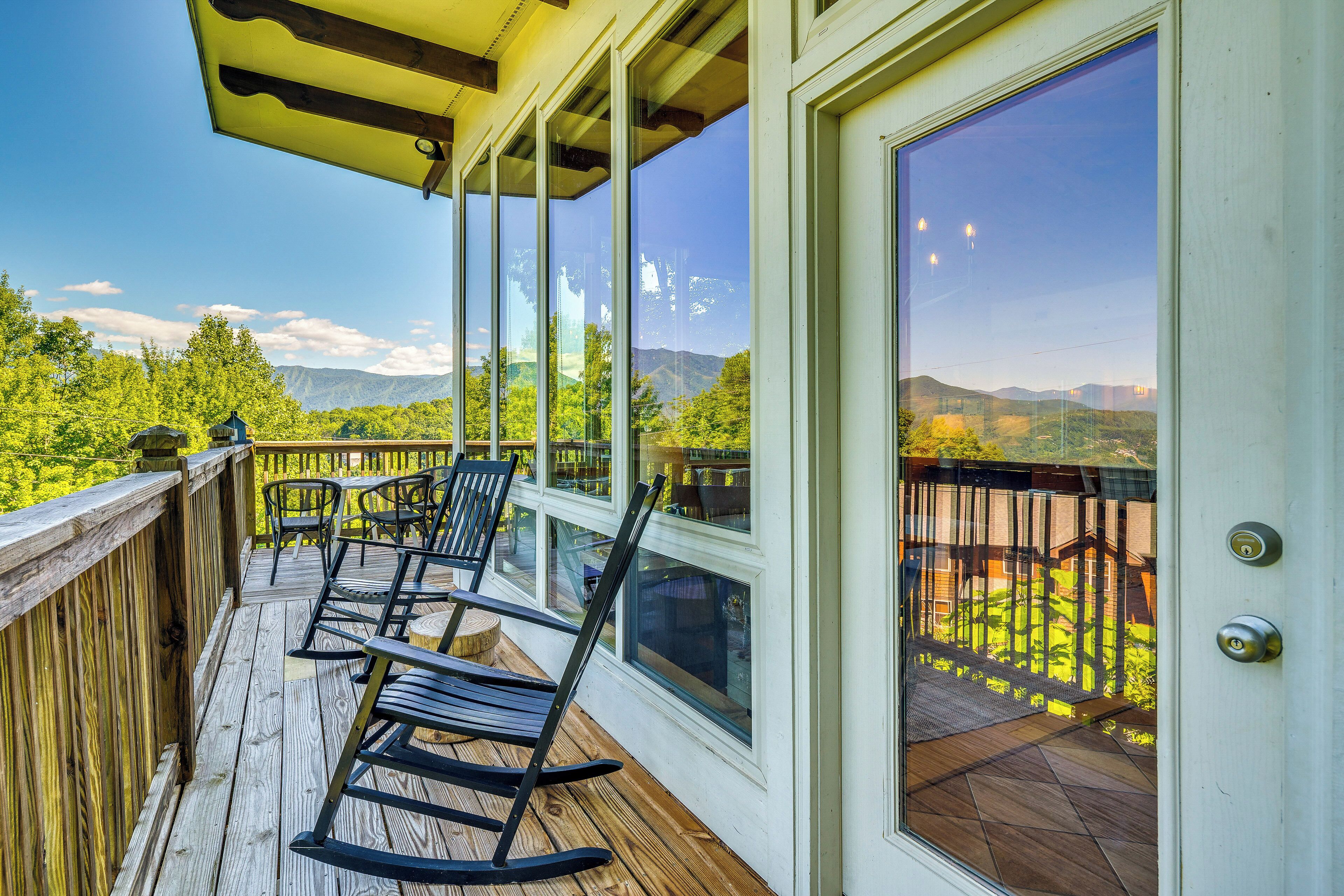Una terraza de madera con mecedoras, una puerta de cristal y vistas a montañas y follaje.