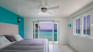 Une chambre avec un lit, un ventilateur de plafond, une vue sur l’océan par une porte coulissante et une fenêtre donnant également sur l’océan.