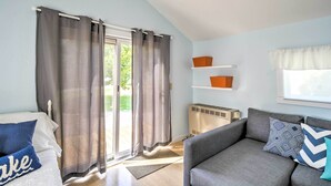 Une pièce comprenant un lit, un canapé gris, une fenêtre avec des rideaux, un radiateur et une étagère avec des boîtes de rangement.