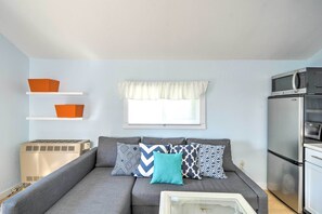 Um espaço de estar compacto com sofá cinza, almofadas estampadas, janela com cortina branca e uma cozinha compacta com micro-ondas e frigorífico.