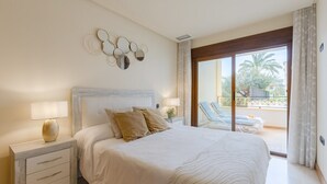 A bedroom with a bed, a nightstand, a lamp, a wall decoration, and a view of a balcony with chairs. - Fidalsa Guardamar Resort (Guardamar)