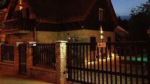 A rustic wooden house with a stone foundation, a gated entrance, and a visible swimming pool in the background.