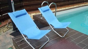 Two blue lounge chairs by a swimming pool with a stone-tiled deck and a wooden fence in the background.