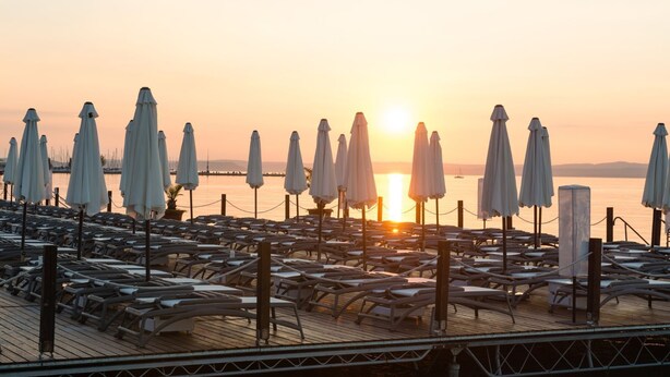 A sunlit deck with rows of lounge chairs and umbrellas, overlooking a calm body of water.