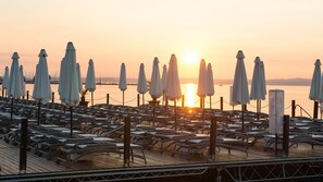A sunlit deck with rows of lounge chairs and umbrellas, overlooking a calm body of water.