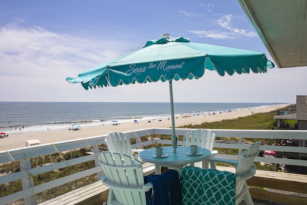 A balcony with a table and chairs, overlooking a beach with umbrellas and people.