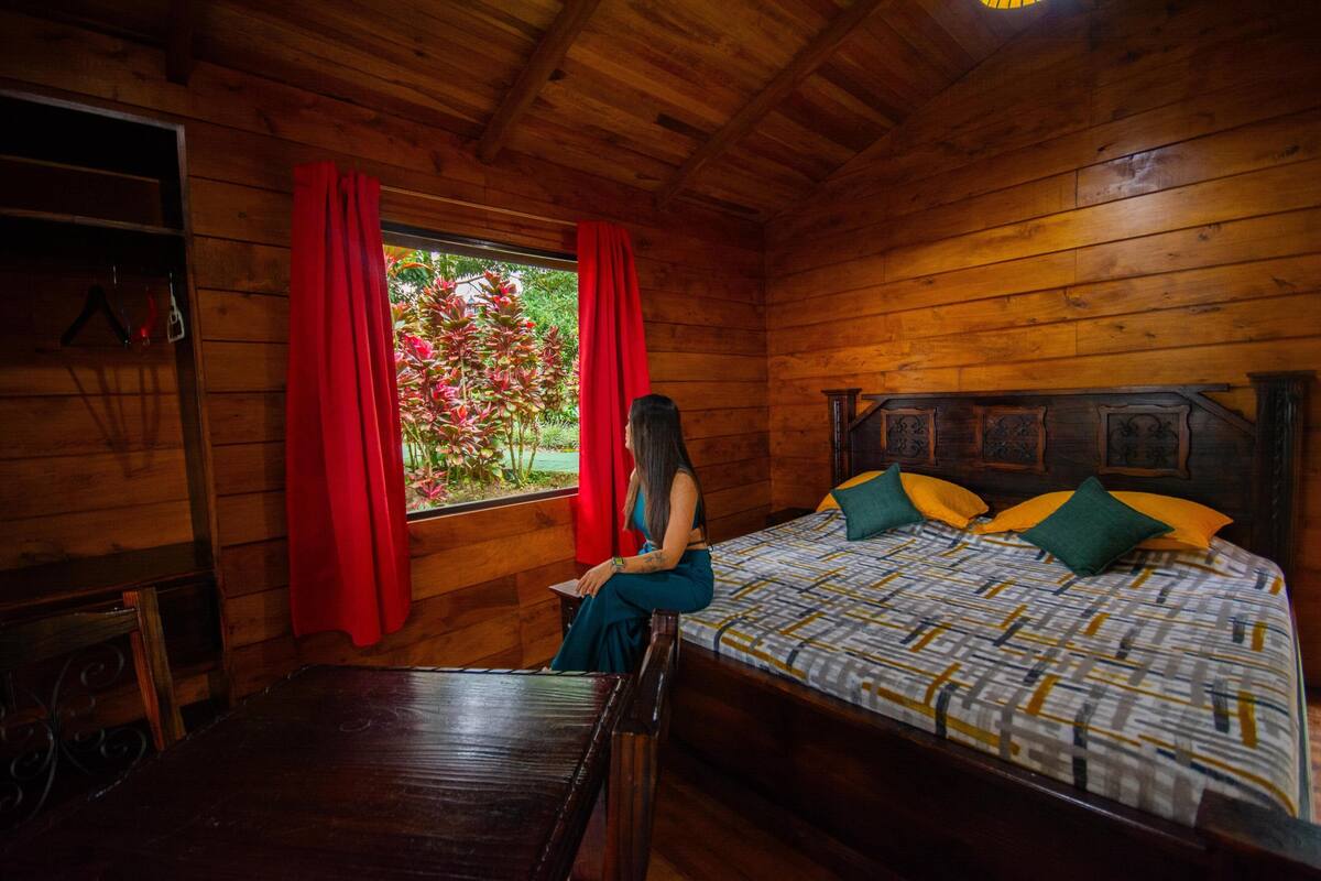 Habitación de cabaña de madera con una cama, una ventana con cortinas rojas y vistas a la vegetación.