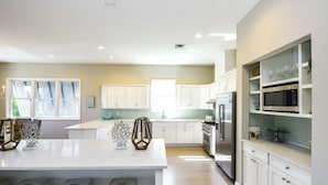 A modern kitchen with white cabinets, a central island with stools, a built-in microwave, and a stainless steel refrigerator. - Canal front Home with Private Dock, Pool, Games, and Water Adventures (Anna Maria)