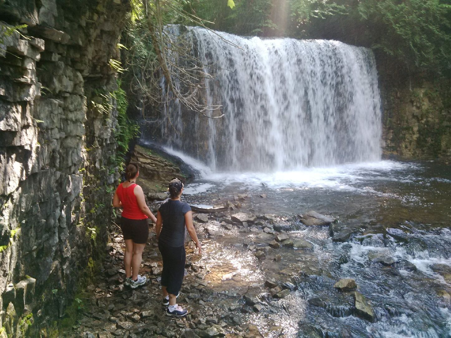 Una cascata in un ambiente naturale, con terreno roccioso e due persone che la osservano.